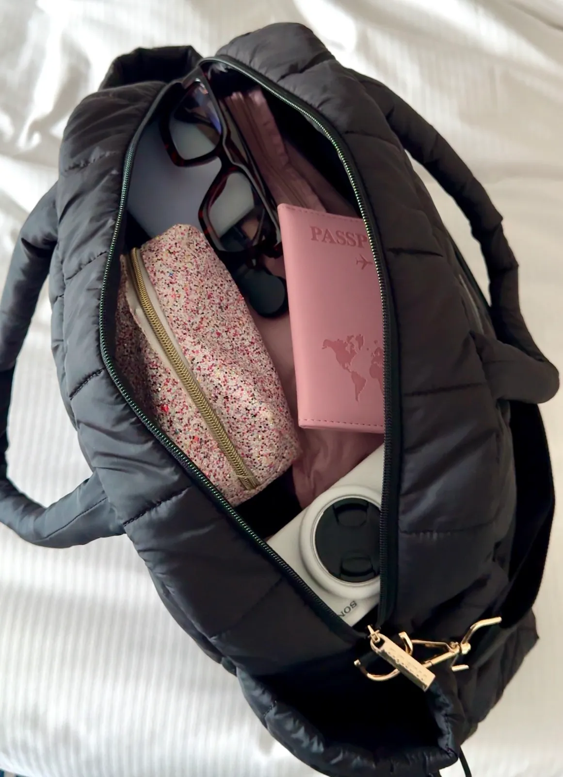 An open black Augustnoa backpack packed with glasses, a pink passport holder, a glitter pouch and a camera