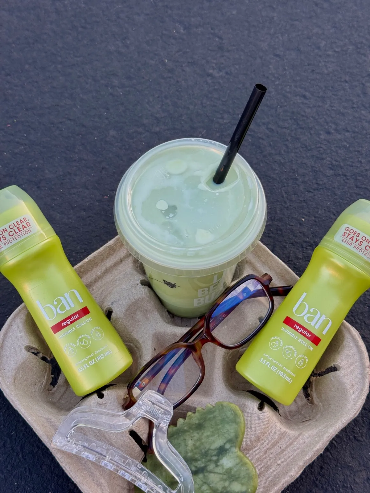 Two Ban roll-on deodorant sticks and an iced green drink in a cardboard cup carrier, with a pair of glasses resting between them