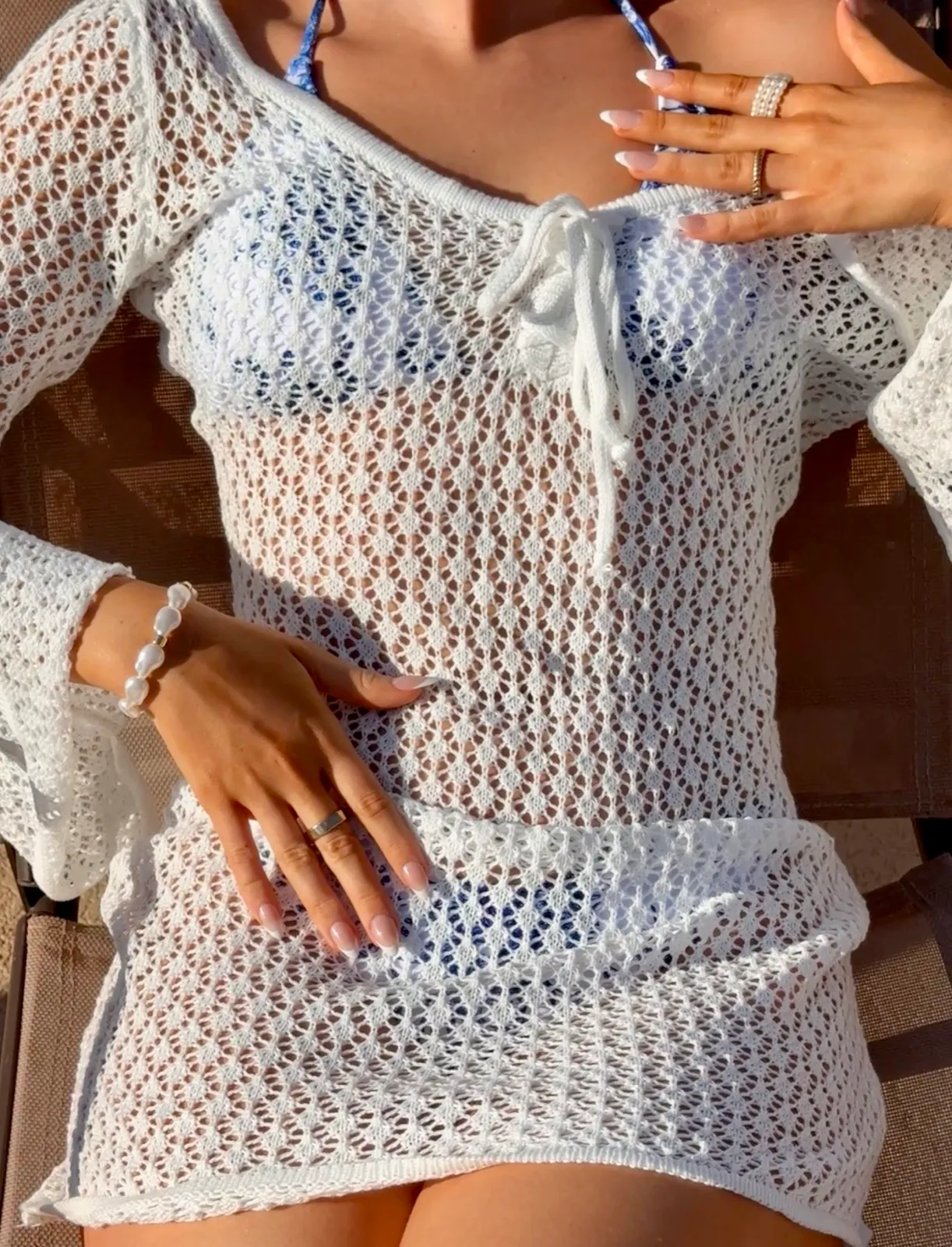 Close-up of a woman in a white crochet cover-up over a blue bikini, hands adjusting the tie at the chest