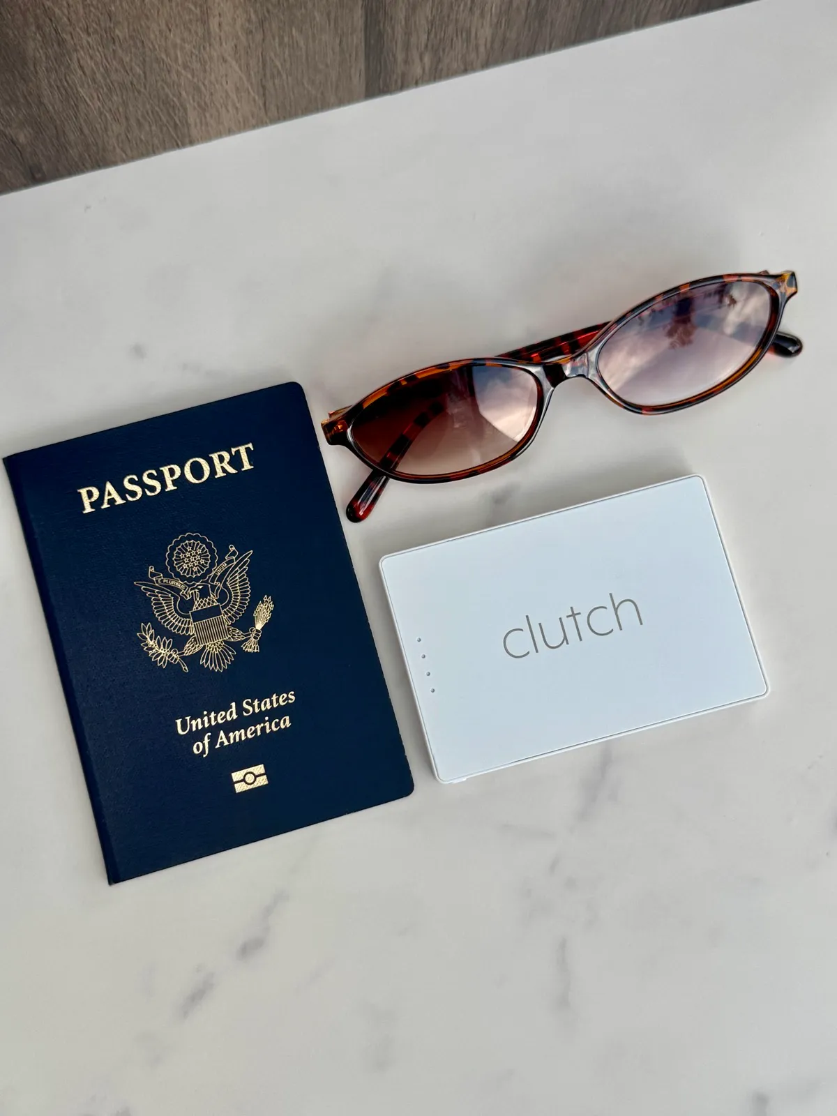 A navy passport, tortoiseshell sunglasses and a white Clutch portable charger arranged on a marble surface