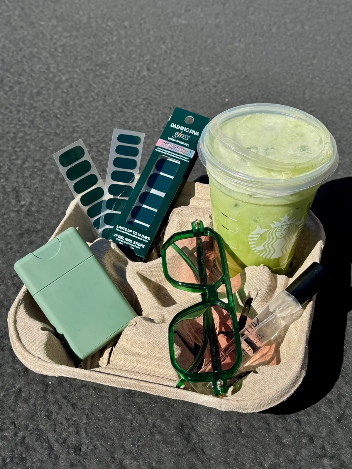 Dashing Diva dark green gel nail strips in their box, an iced matcha drink and green sunglasses arranged in a cardboard cup carrier