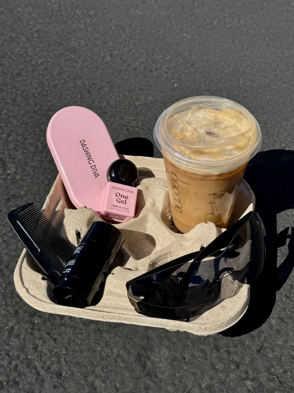 A pink Dashing Diva case, a bottle of One Gel polish, a comb and sunglasses beside an iced coffee in a cardboard cup carrier