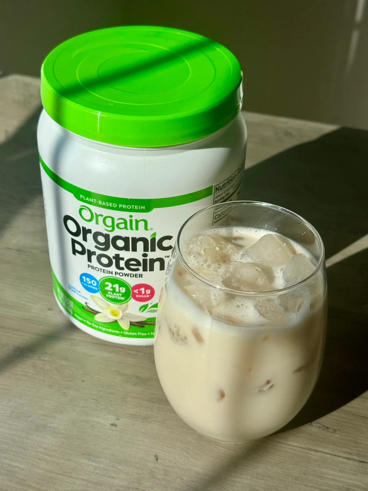 A tub of Orgain Organic Protein powder beside a glass of iced protein shake