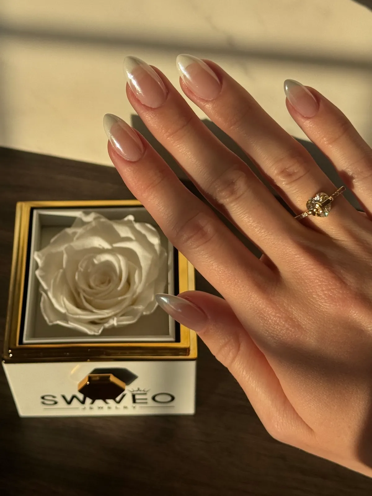 A hand with french tip nails and a diamond ring beside a Swaveo Jewelry box holding a white preserved rose